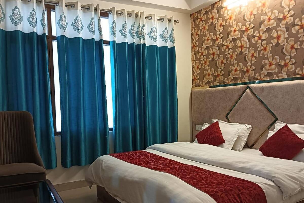 A hotel room with a bed, two red pillows, a brown chair, and a window with blue curtains.