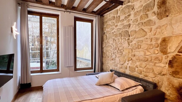 Ein Schlafzimmer mit Steinwand, Holzbalken, einem Bett mit Sitzbank und einem Fernseher.