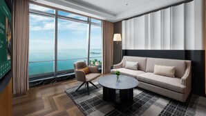 A modern hotel room with a large window offering a view of the sea, a sofa, a chair, and a round coffee table. - MaxX Harbour-view Qingdao (Qingdao)