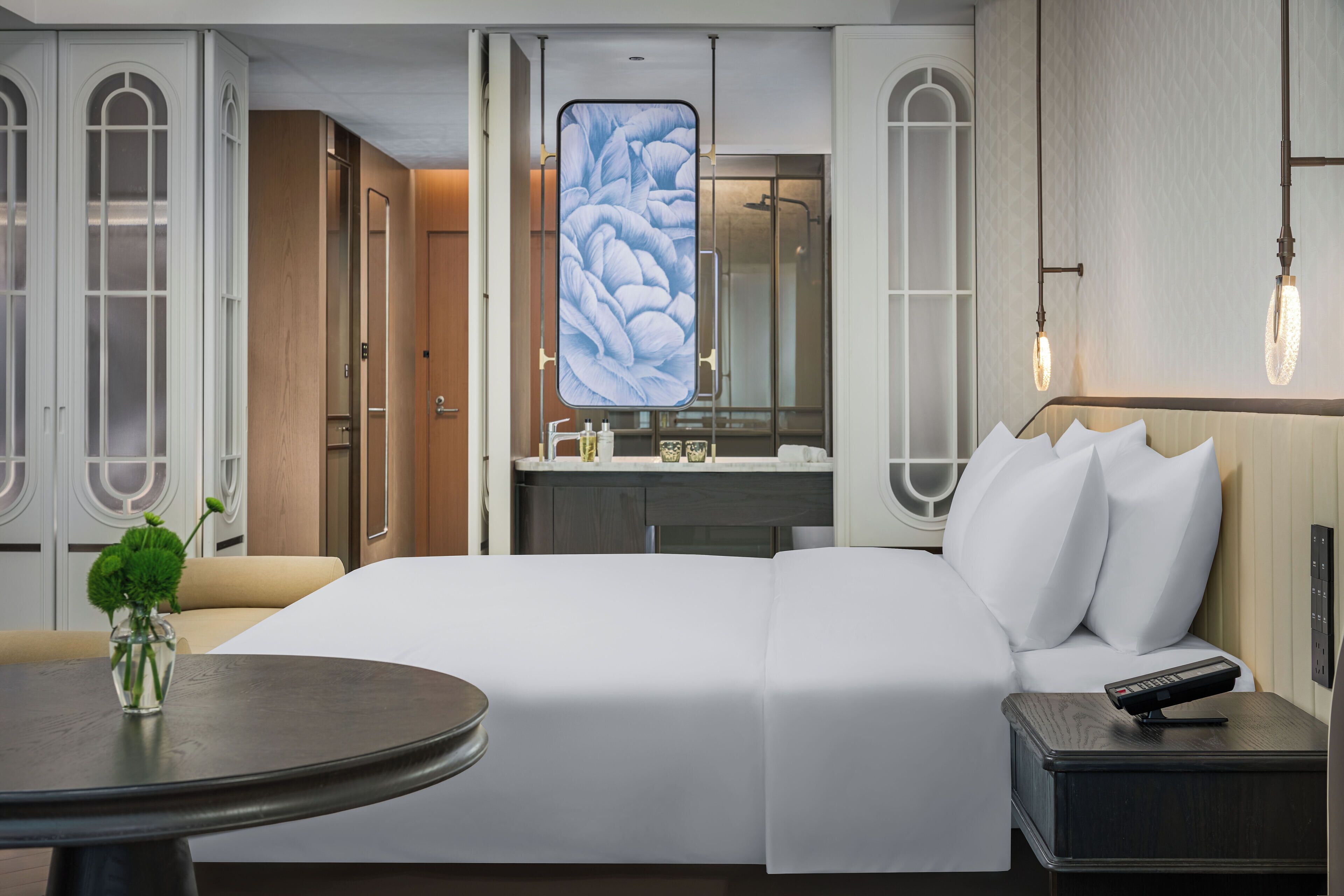Une chambre d’hôtel moderne avec un grand lit, une table de chevet, une table ronde ornée d’un vase et un grand miroir décoré de motifs floraux.