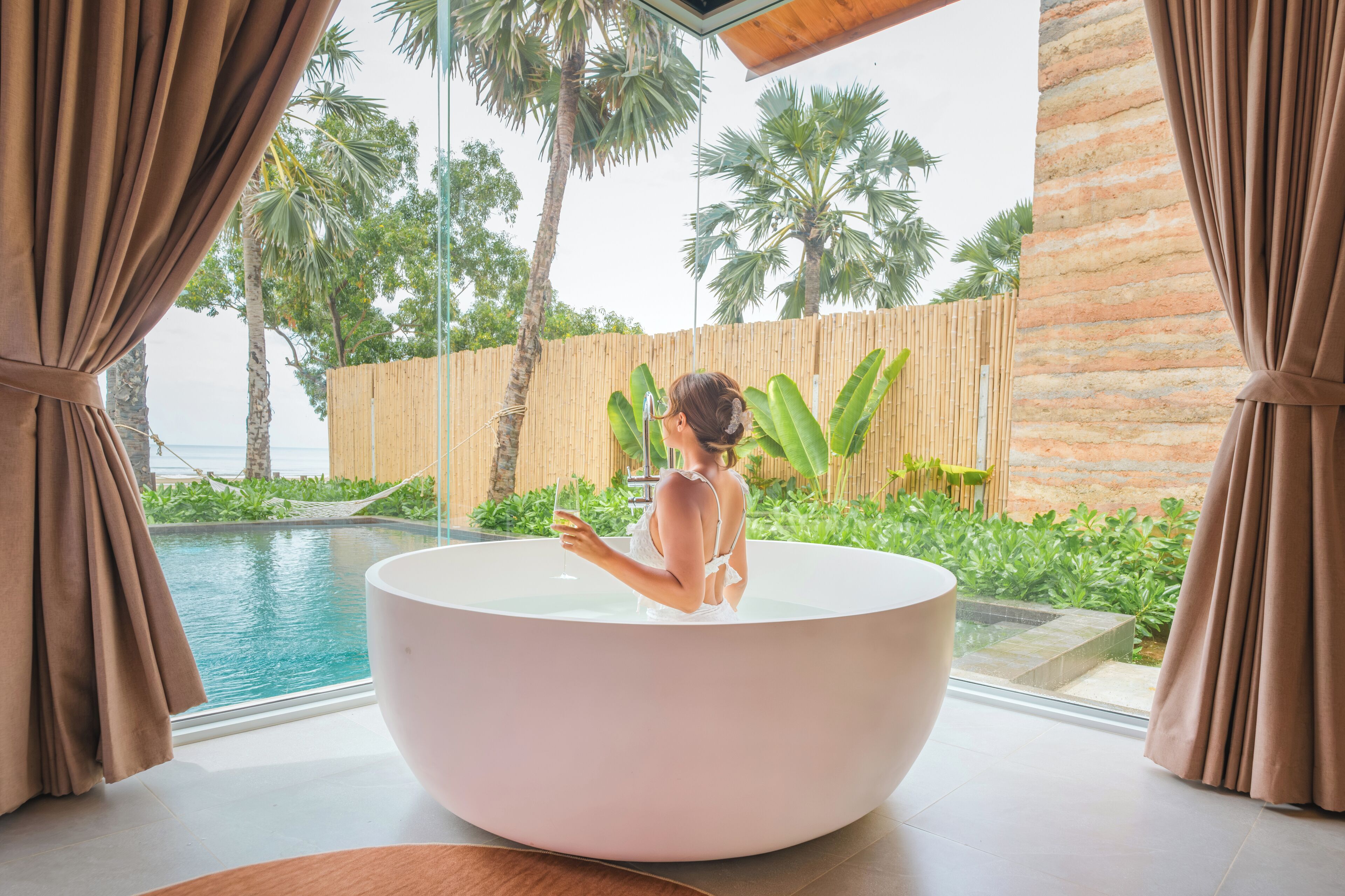 Eine Frau in einer großen Badewanne mit Blick auf einen Pool und Palmen.