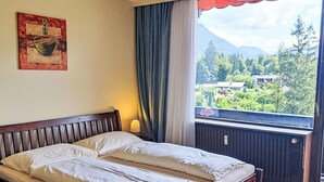 A double bed with a wooden headboard, a bedside lamp, a window with a view of greenery and mountains, and a painting on the wall. - Wohnung 414 Bergblick by Interhome (Strobl)