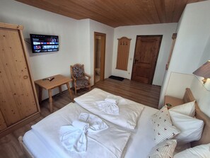 A hotel room with a wooden ceiling, a TV mounted on the wall, a desk with a chair, and a bed with neatly made white linens. - Pension Razil (Ladis)
