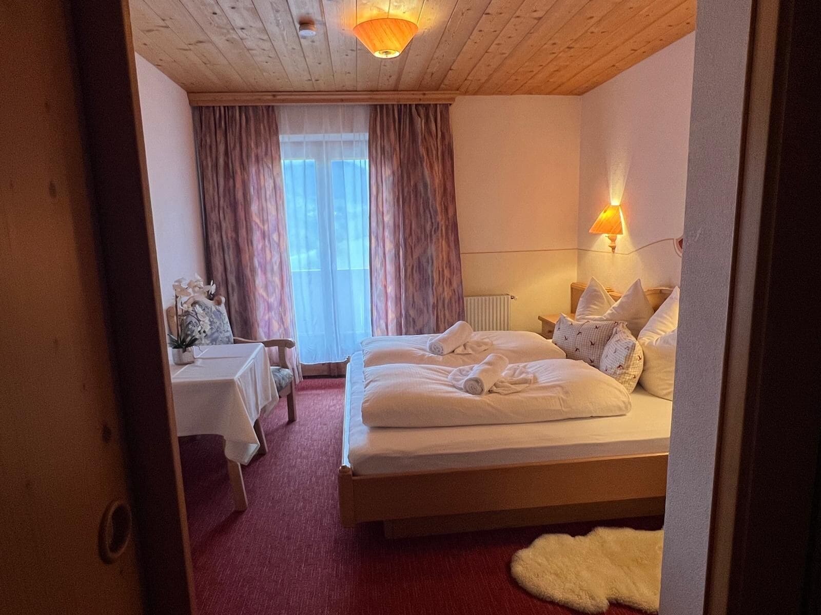 A hotel room with a large bed, a small table with a chair, and a window with curtains.