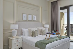 A neatly arranged hotel room with a large bed, bedside lamps, a small table with a book and a bowl of fruit, and a balcony with a view. - Maison Privee - Creekside Elegance w/ Canal Vws at Creek Beach (Dubai)