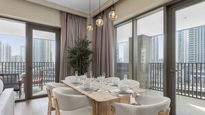 A modern dining area with a wooden table set for six, surrounded by plush chairs, a large potted plant, and a view of high-rise buildings through large windows. - Maison Privee - Creekside Elegance w/ Canal Vws at Creek Beach (Dubai)