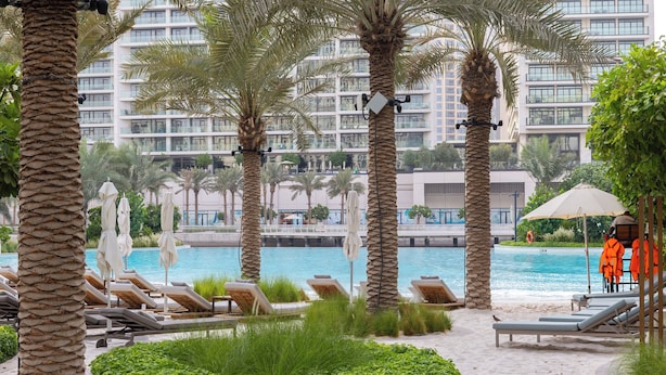A poolside area with lounge chairs, palm trees, and a view of a high-rise building. - Maison Privee - Creekside Elegance w/ Canal Vws at Creek Beach (Dubai)
