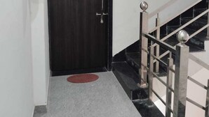 A hallway with a dark wooden door, a red fire extinguisher, a green mat, and a staircase with metal railings. - Shree Kalyan Palace (Danta Ramgarh)