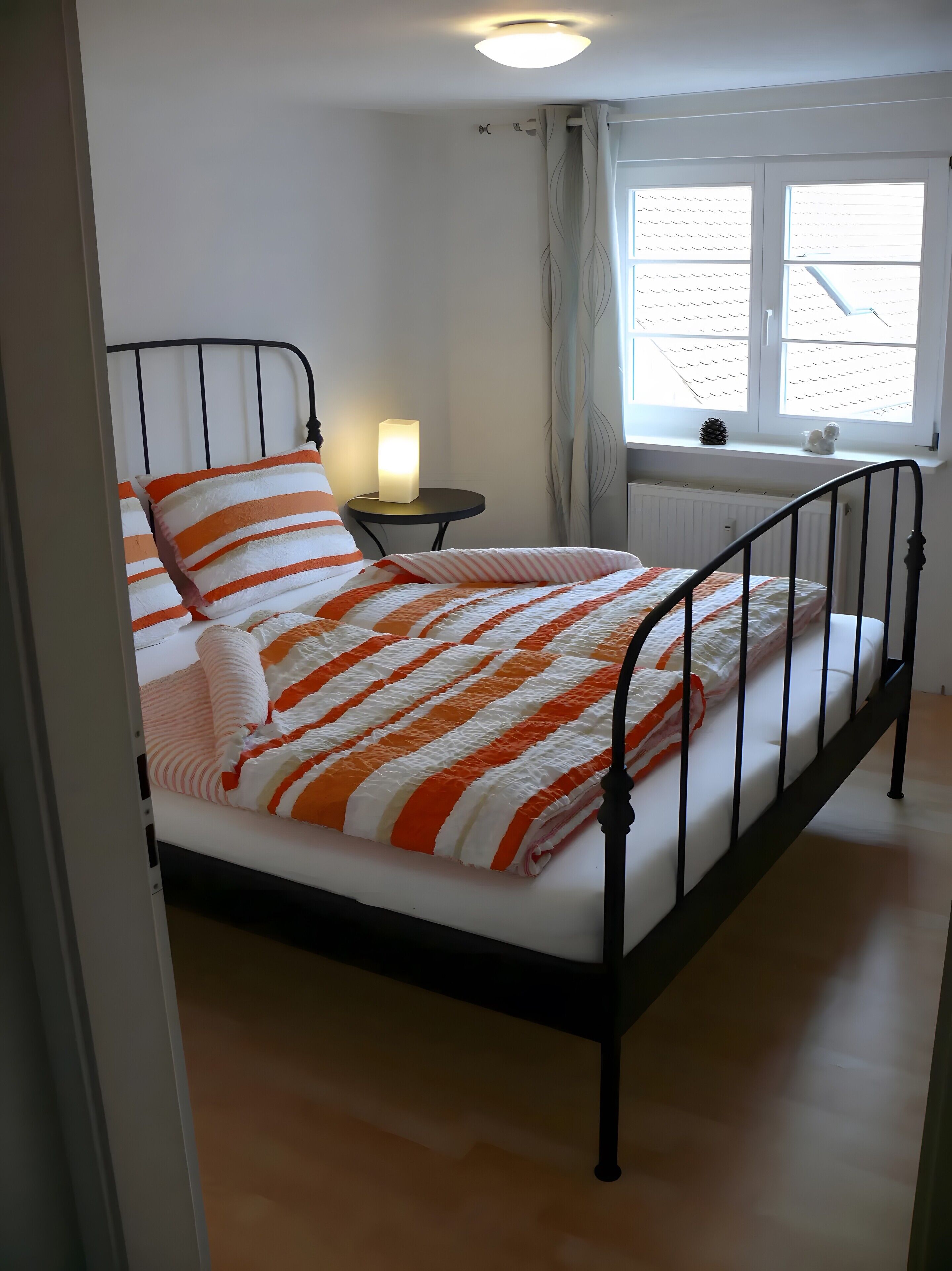 Una cama con una manta a rayas, una mesita de noche con una lámpara, una ventana con cortinas y un radiador.