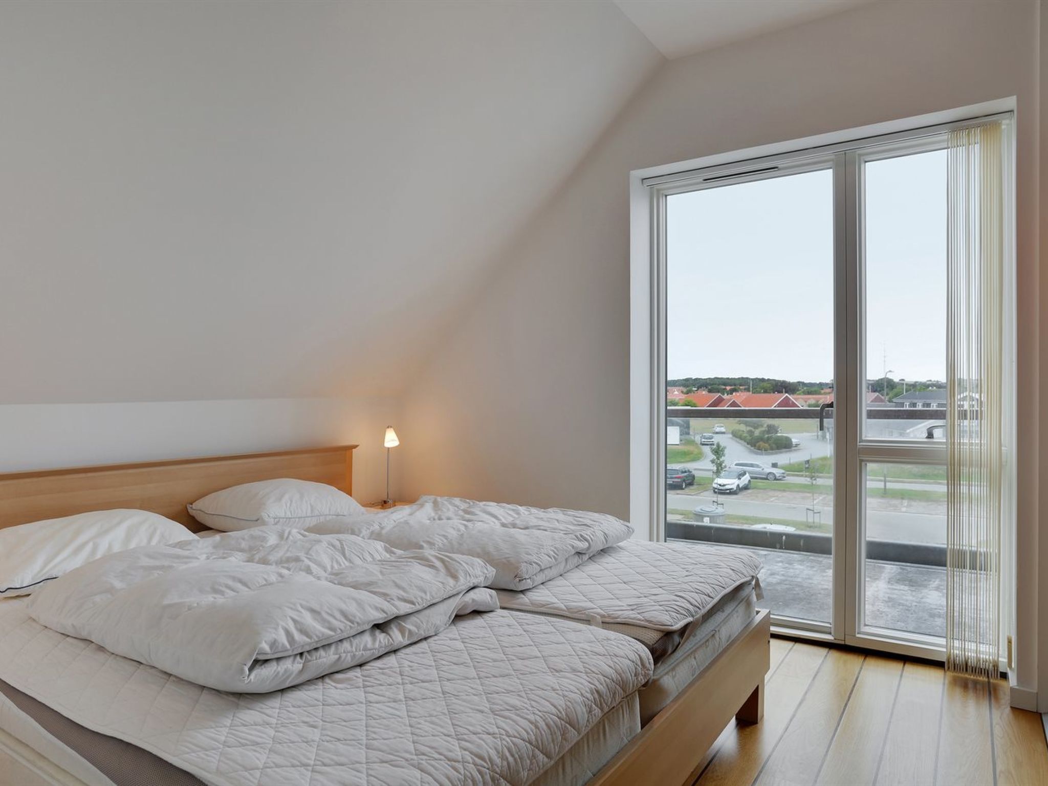 Et soveværelse med en seng, et natbord og et vindue med udsigt til en parkeringsplads og bygninger.