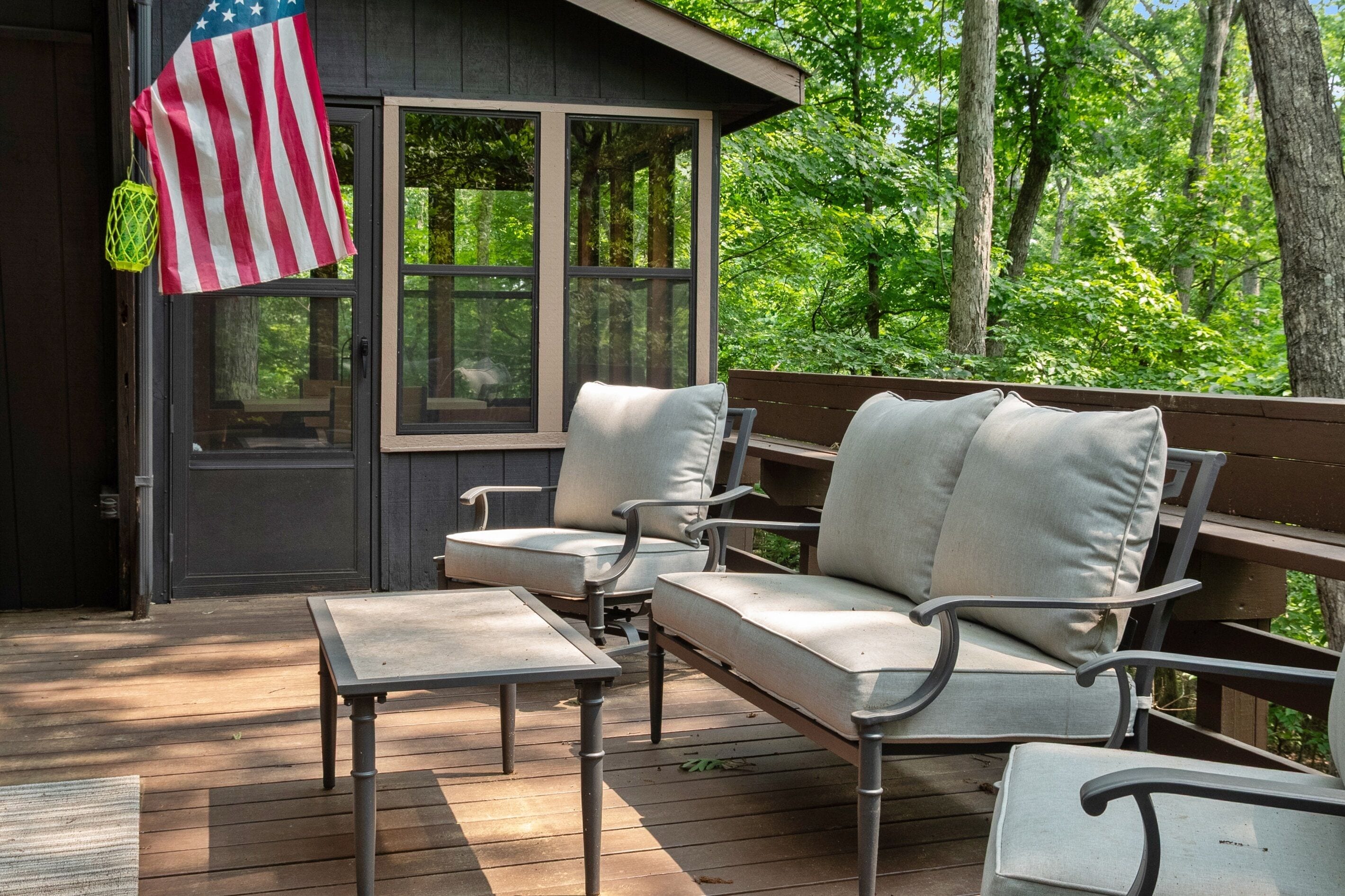 Een houten terras met een bank, een tafel, een Amerikaanse vlag en uitzicht op bomen.