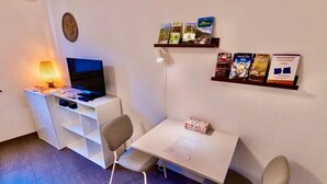 A small room with a desk, a chair, a television, a lamp, and a shelf with brochures.
