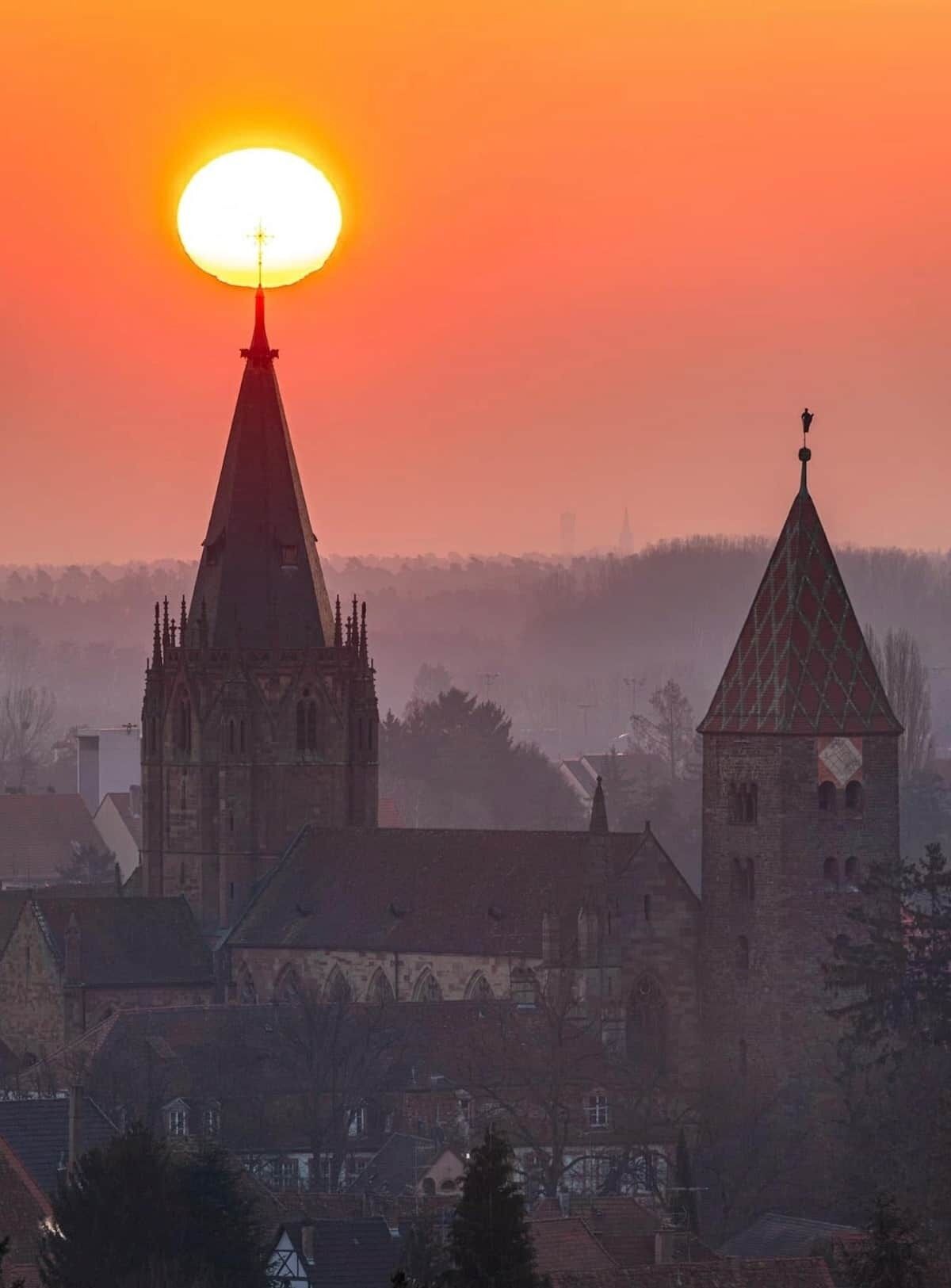 Een kerk met een opvallende torenspits en een kleinere aanwezige toren, voor een achtergrond van een zonsondergang.
