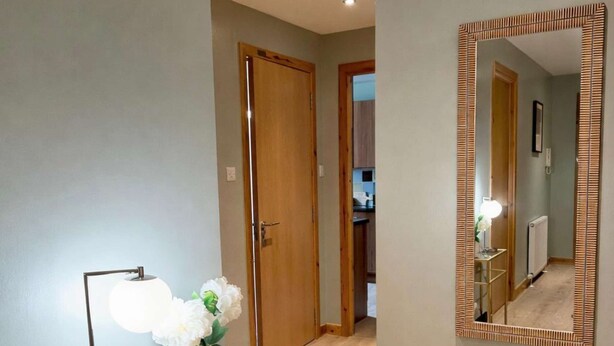 A hallway with a wooden floor, a mirror with a striped frame, a vase with flowers, and a floor lamp.