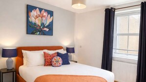 A bedroom with a bed, two bedside tables, a lamp, a painting, and a window with curtains.