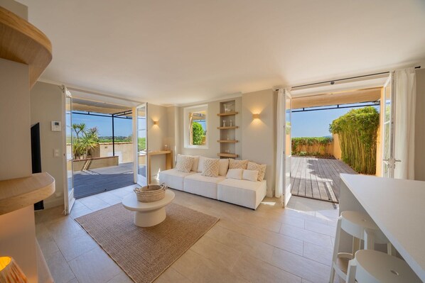 A modern living room with a white sofa, a coffee table, and a dining area with white chairs. There are built-in shelves and a large window offering a view of the outdoors.