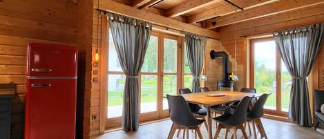 A wooden interior with a dining table, chairs, a red refrigerator, and large windows with curtains.