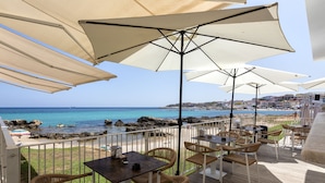 Una terraza con sillones y mesas de mimbre, con vistas a una playa y un litoral rocoso.