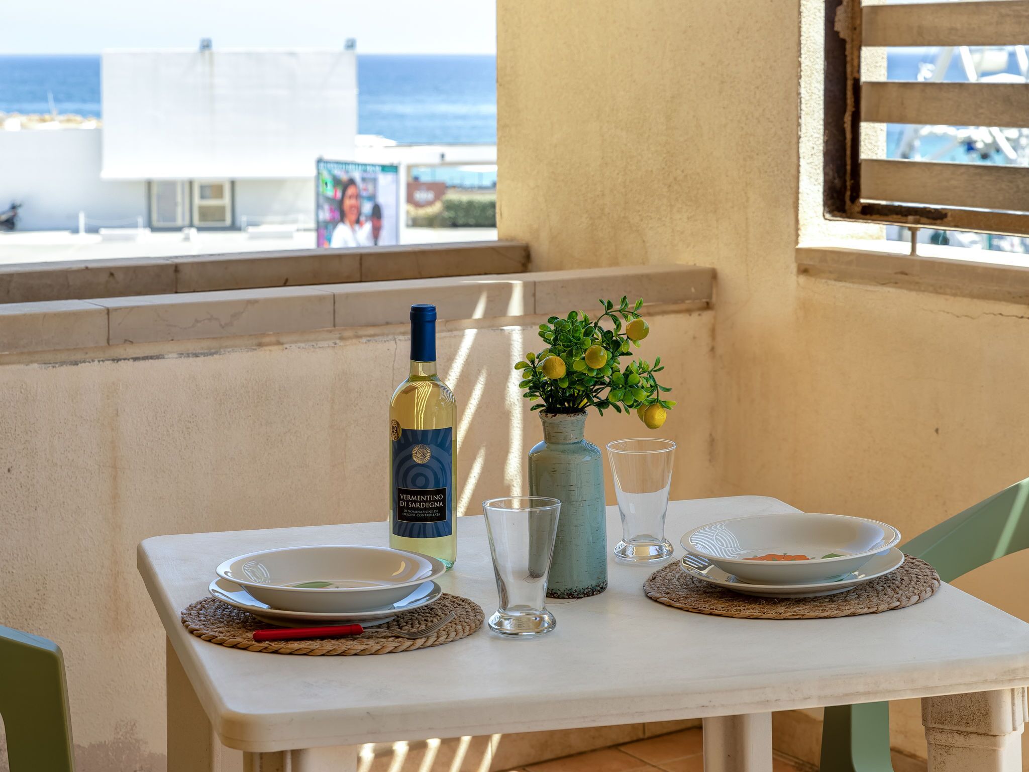 A table set with plates, a bottle, a vase with lemons, and glasses on a balcony overlooking the sea.