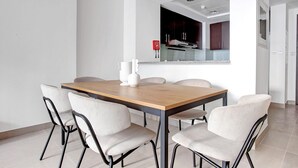 Un comedor moderno con una mesa de madera y seis sillas, una cocina visible a través de un vano abierto, y un detector de incendios en la pared.
