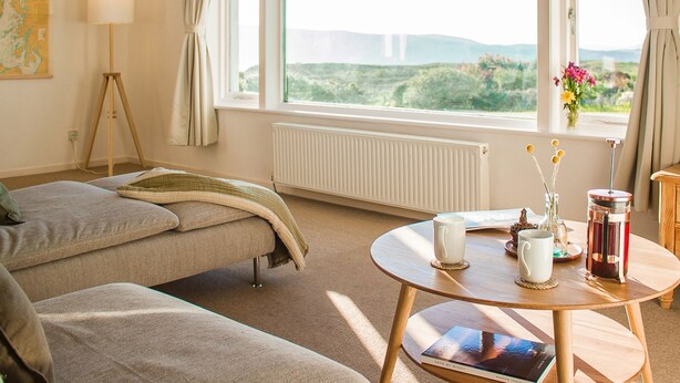 A cozy room with a bed, a small table with a book, a lamp, and a view of the outdoors.