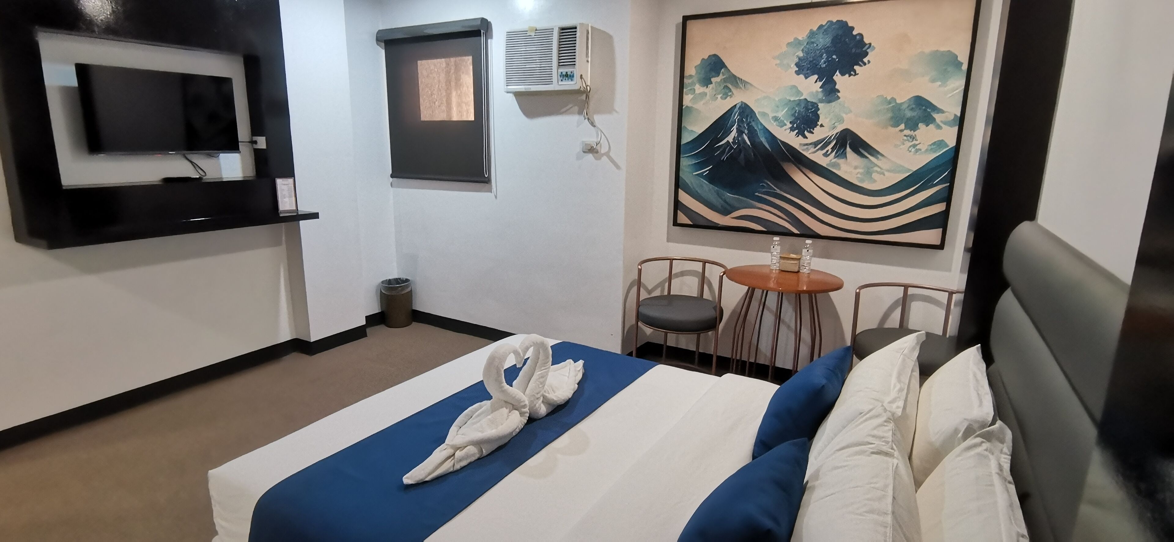 Et hotelværelse med en seng, et tv ophængt på væggen, et maleri, et lille bord med stole og en skraldespand.