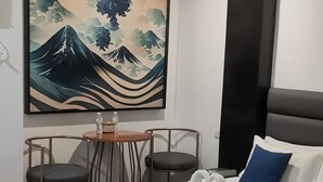 Hotellihuone, jossa on sänky, työpöytä ja kaksi tuolia, suuri taulu seinällä sekä musta pylväs.