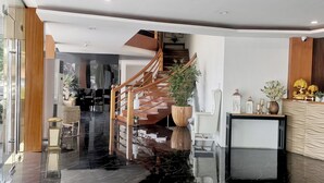 Moderni hotellin aula, jossa on marmorilattia, puinen portaikko, vastaanottotiski ja suuri ruukussa oleva kasvi.