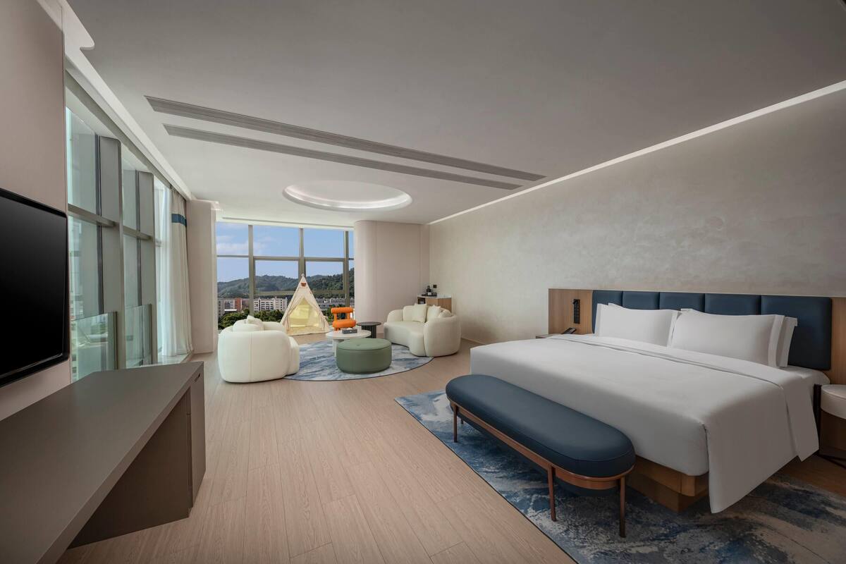 Ein modernes Hotelzimmer mit einem großen Bett, einem Flachbildfernseher, einer Sitzecke mit Sofa und Hocker sowie Blick auf die Stadt durch große Fenster.