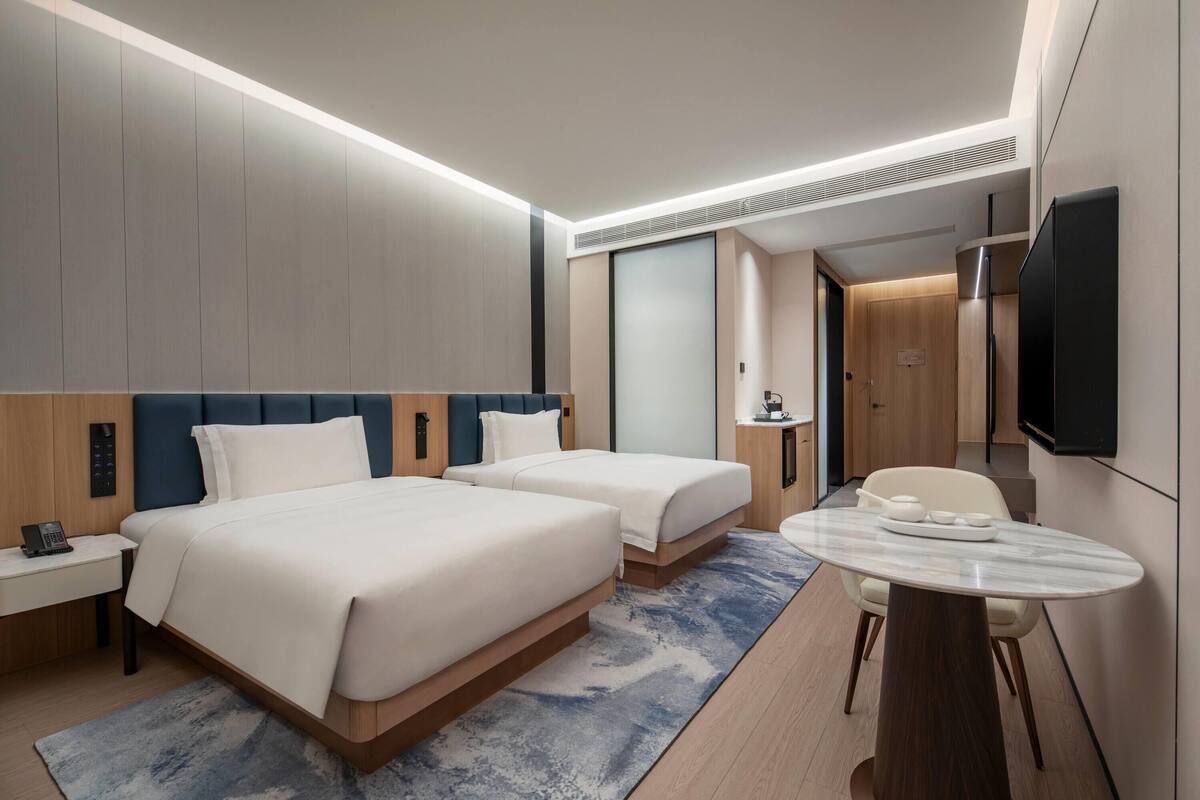 Ein Hotelzimmer mit zwei Betten, einem Schreibtisch, einem Sessel, einem Fernseher und einem kleinen Tischchen mit Tablett.