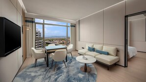 A modern hotel room with a large window offering a city view, a flat-screen TV mounted on the wall, a sofa with blue cushions, a coffee table, and a dining area with chairs and a table. - Days Hotel by Wyndham Zhuhai Jinwan (Zhuhai)