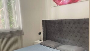 Une chambre à coucher avec une tête de lit capitonnée grise, un lit simple avec des oreillers gris et blancs, une table de chevet blanche avec une lampe, et une fenêtre avec des stores.