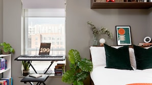 A bedroom with a bed, a desk with a keyboard, a window with a view of buildings, and a bookshelf with books. - For Students Only - Modern Rooms in Lincoln (Lincoln)