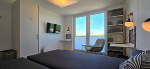 Ein modernes Schlafzimmer mit einem großen Bett, einem Schreibtisch mit Stuhl, einem Fernseher und einem Fenster mit Blick auf den Himmel.