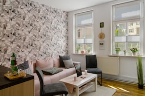 A living room with a floral wallpaper, a sofa, a coffee table, and a chair. There are windows with blinds, a radiator, and a potted plant.