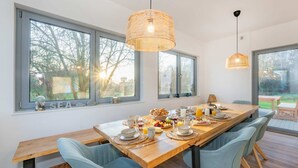 A dining area with a long wooden table, blue chairs, and a view of the outdoors through large windows. - "Haffhus" Vacation Home with a Balcony, Terrace, and 2 Bathrooms - Modern Vacation Home (Usedom)