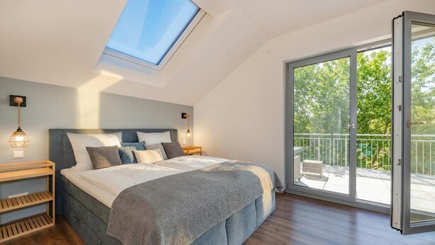 A bedroom with a skylight, a bed with a grey blanket, wooden nightstands, and a view of a balcony. - "Haffhus" Vacation Home with a Balcony, Terrace, and 2 Bathrooms - Modern Vacation Home (Usedom)