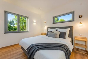 A bedroom with a bed, two bedside tables, a nightstand, and two windows with views of greenery. - "Haffhus" Vacation Home with a Balcony, Terrace, and 2 Bathrooms - Modern Vacation Home (Usedom)