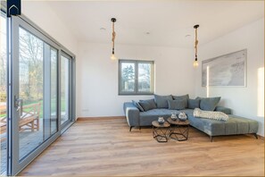 Un salon moderne avec un canapé d’angle gris, deux lampes suspendues, une table basse et une grande fenêtre donnant sur l’extérieur.