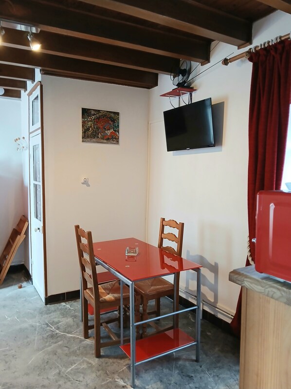 Una habitación pequeña con una mesa roja y dos sillas de madera, un televisor montado en la pared y una cortina roja.