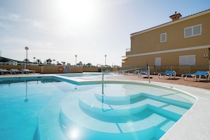 A swimming pool with a clear water design, surrounded by lounge chairs and a building with a tiled roof. - LOMA ESTRELLA (Arguineguín)