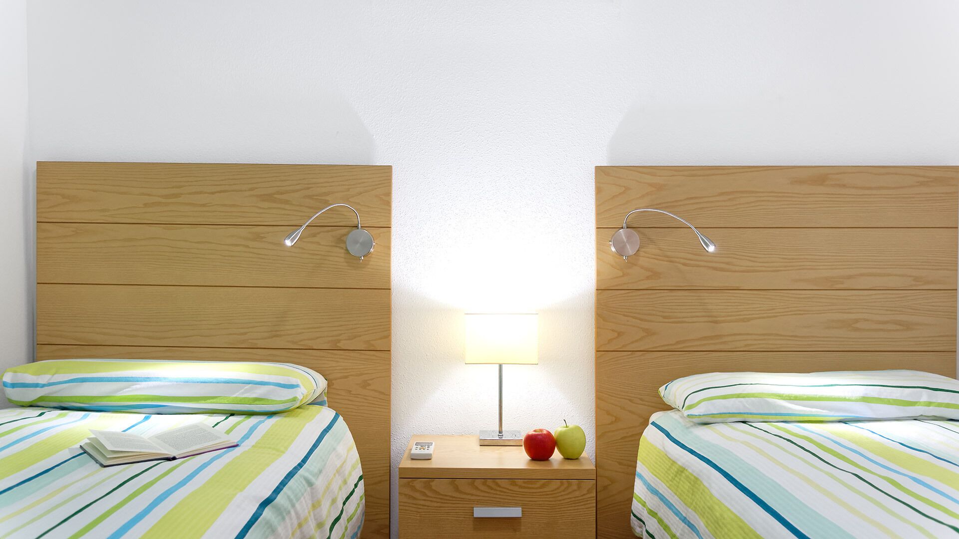 Een hotelkamer met twee enkele bedden, een nachtkastje met een lamp, en een bijzettafeltje met een appel en een boek.