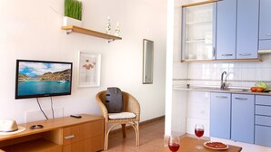 Een ruimtelijk ingerichte zithoek met een televisie, een rieten stoel, een klein bureau, een keuken met blauwe kastjes en een eettafel met een bord eten en een glas rode wijn.