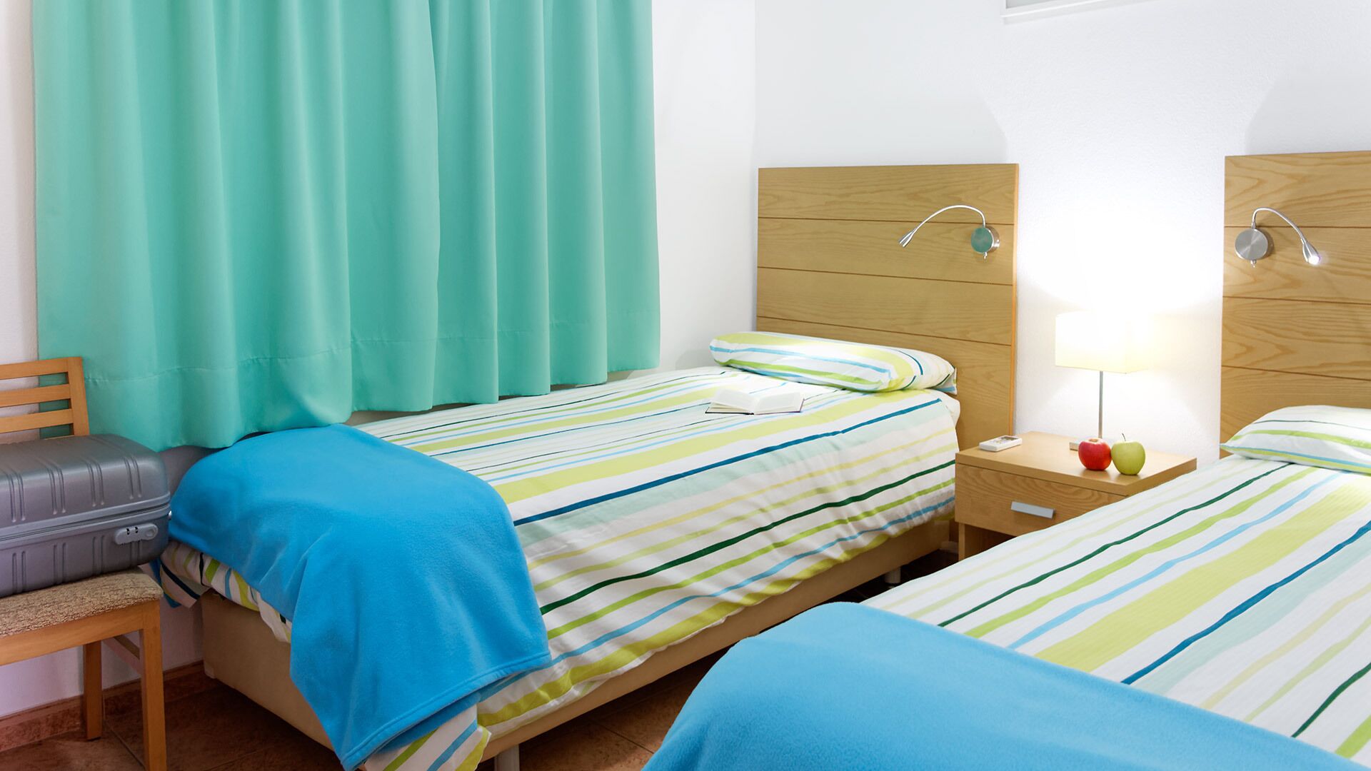 Habitació d'hotel amb dos llits, un capçal de fusta, una tauleta de nit amb una làmpada i una maleta sobre una cadira.