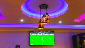 A room with a flat-screen TV mounted on the wall, a ceiling with circular lighting, and a hanging pendant light fixture. - Holiday Premier Hotel (Ol Kalou)