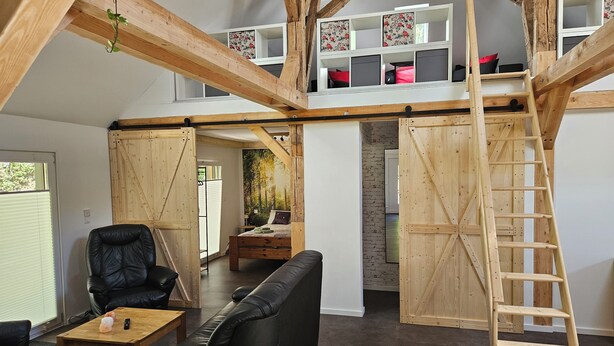 A loft-style bedroom with exposed wooden beams, a loft bed, a wooden ladder, a black leather sofa, a wooden coffee table, and a large window.