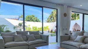 A modern living room with two beige sofas, a wooden coffee table, and large windows offering a view of a white building and greenery.