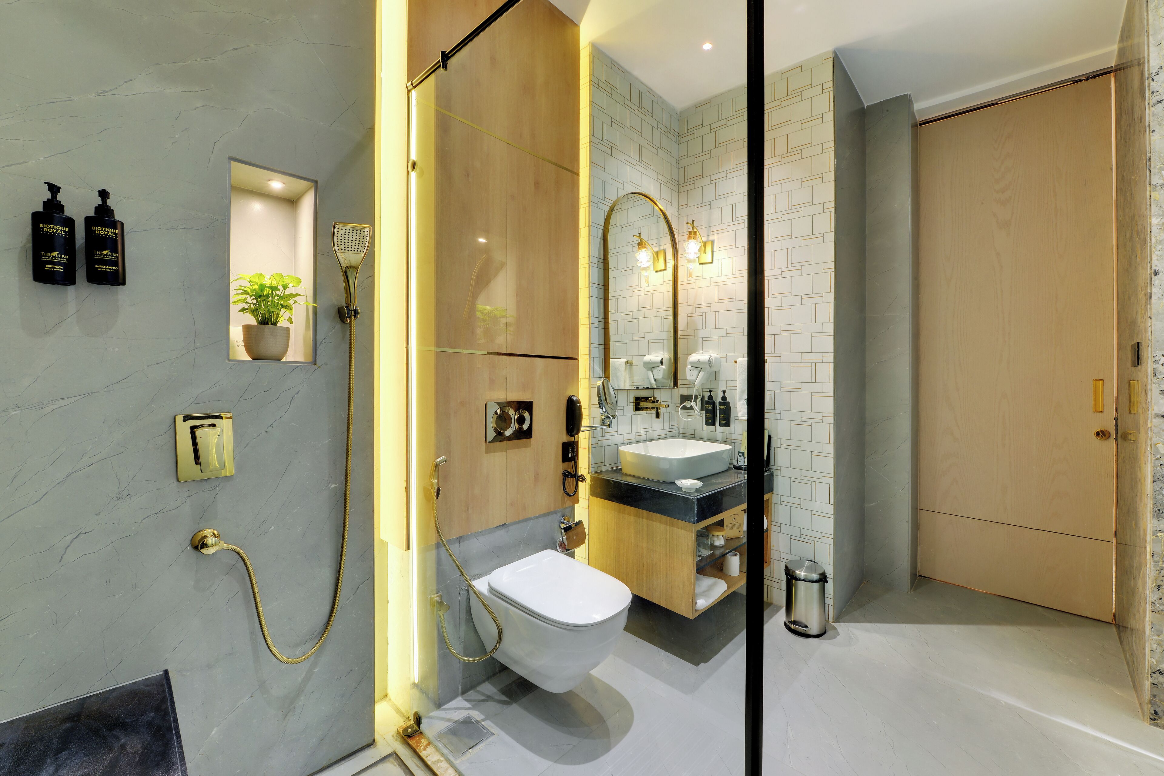 Un baño moderno con inodoro, lavabo y ducha. Hay dos botellas de champú en la pared, una planta en maceta y un cubo de basura.