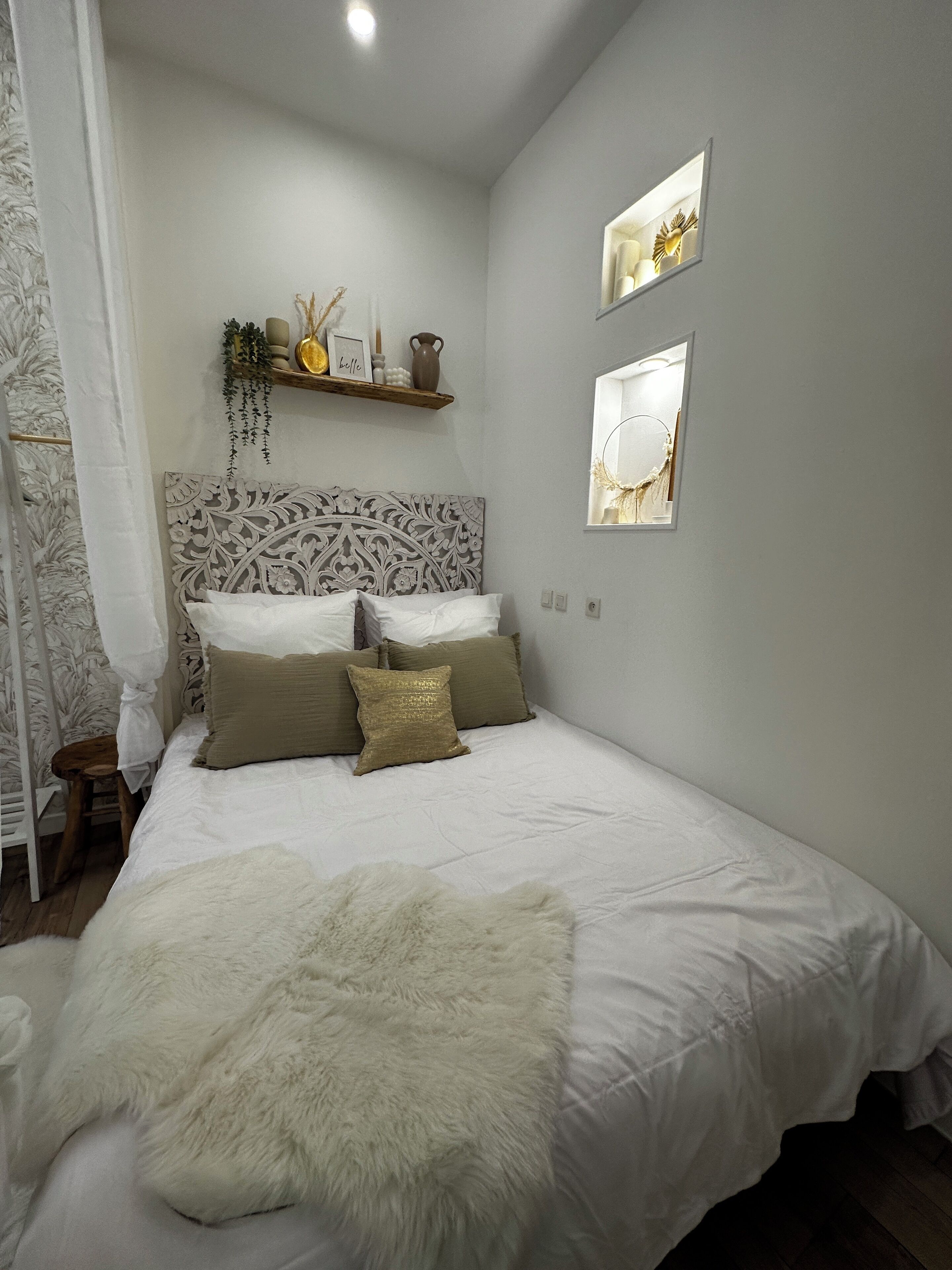 A bedroom with a large bed, a decorative headboard, a small stool, and a shelf with decorative items.