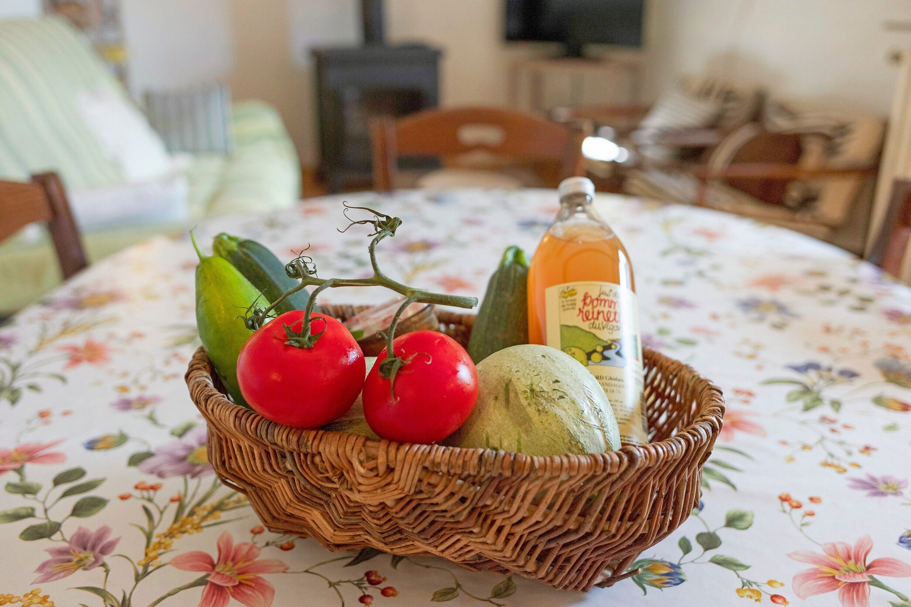 Un panier en osier contenant des tomates, un concombre et une bouteille de jus, posé sur une nappe à motifs floraux.